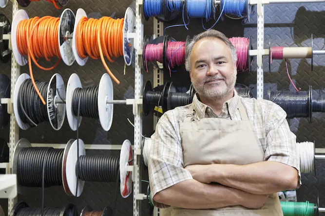 Man with gray hair and waring an apron is standing in front of a wall of electrical wire.